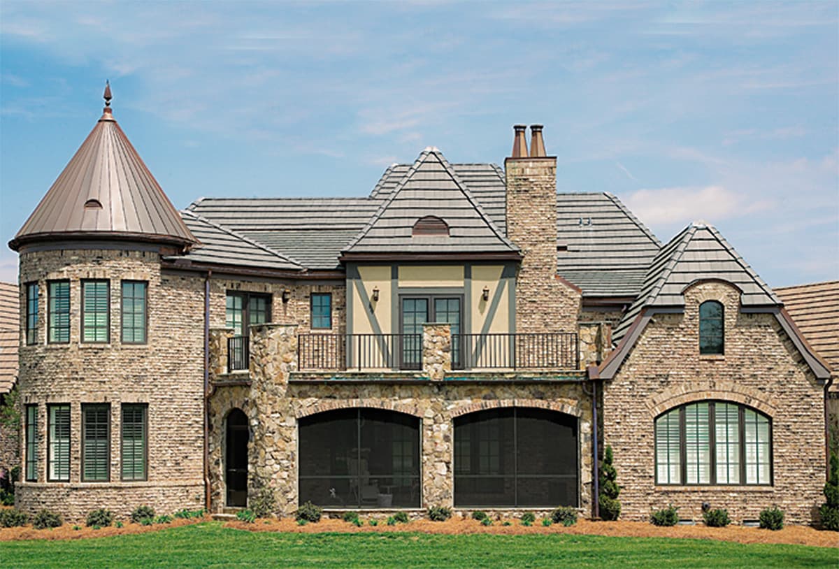 House plan exterior with stone and brick facade, prominent turret with copper roof, multiple gables, and arched screened openings.