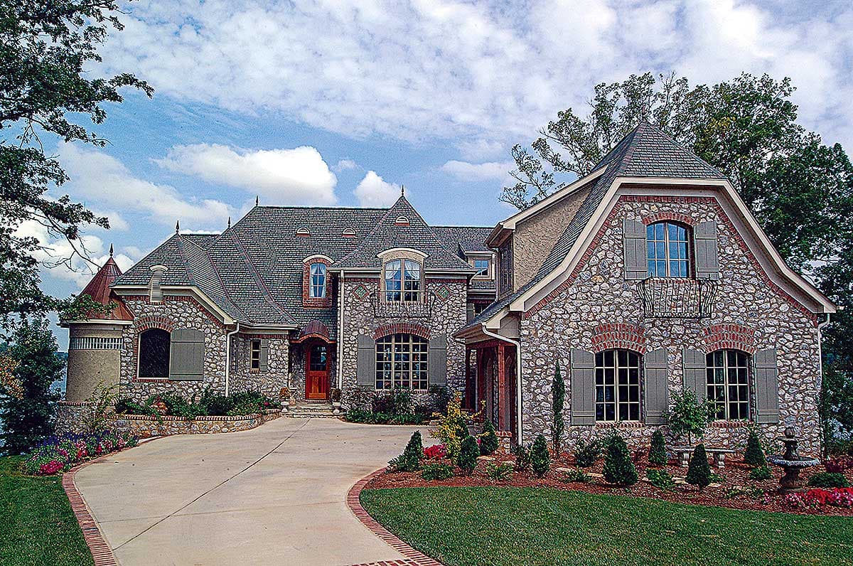 House plan exterior. Stone facade with multiple gables and a multi-level roofline. Features include arched windows and a brick-lined driveway.