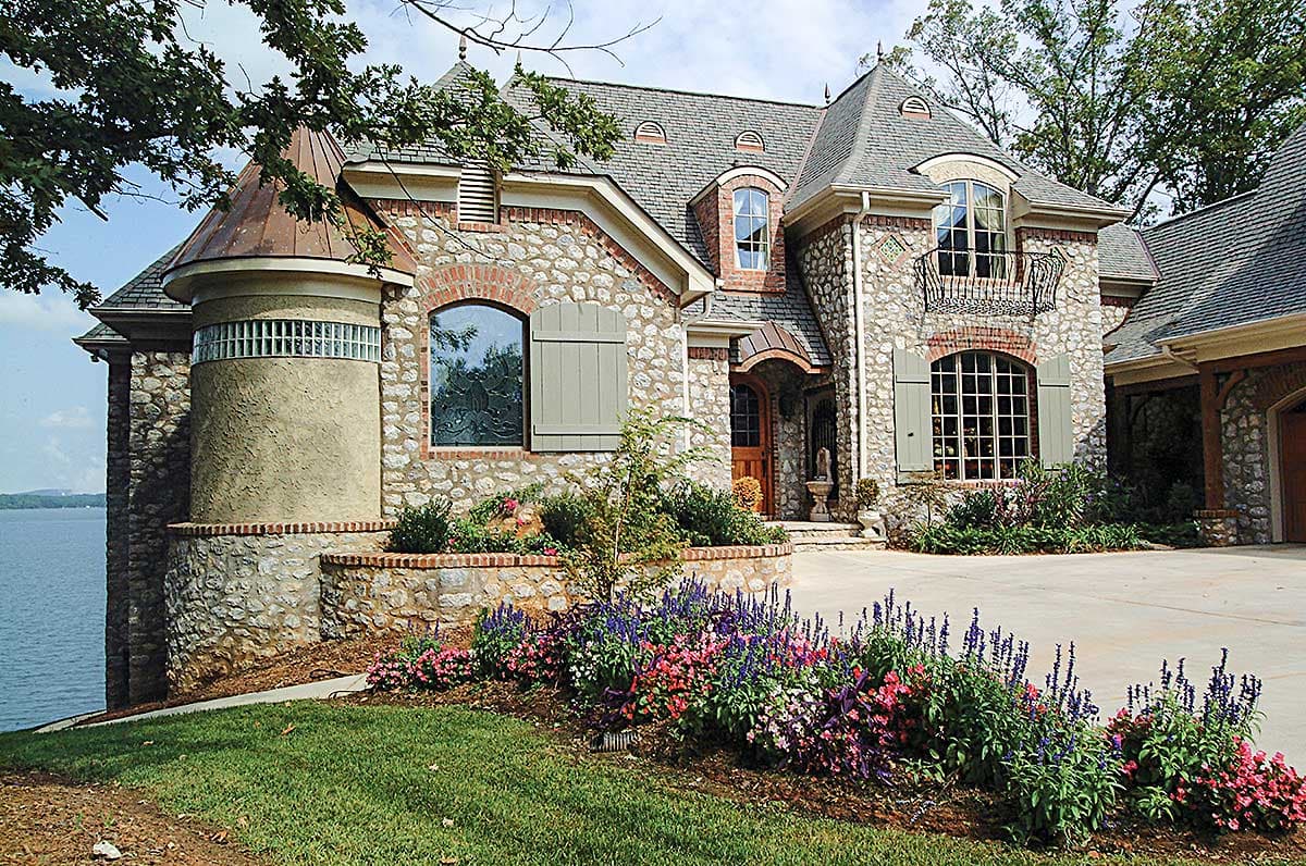House plan exterior with stone facade, turret, mansard roof, arched windows, and decorative balcony.