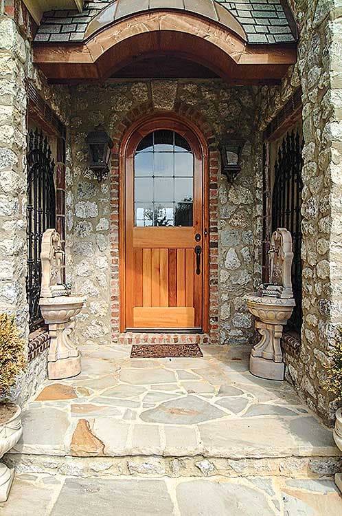 Stone and brick exterior with arched wooden door, leaded glass window, copper roof detail, and two small fountains.