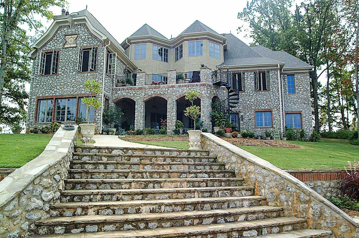 House plan exterior: multi-story stone residence with arched loggia, balconies, spiral staircase, and prominent front steps.