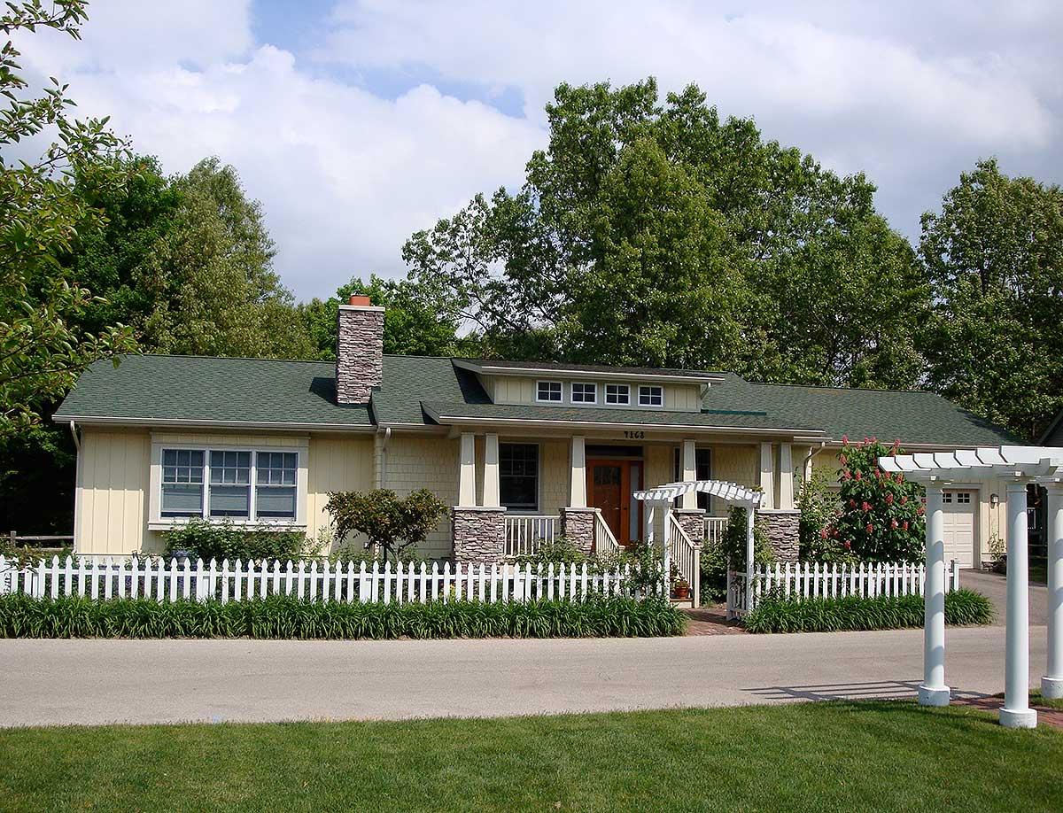 Ranch house plan exterior with green roof, stone chimney, covered entry porch, and white picket fence.