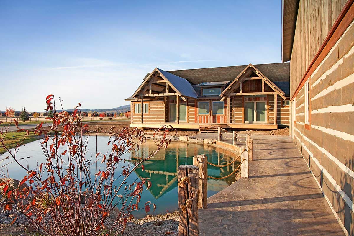 Log house plan exterior with prominent gables, exposed timber framing, and a wide wraparound deck next to a pond.