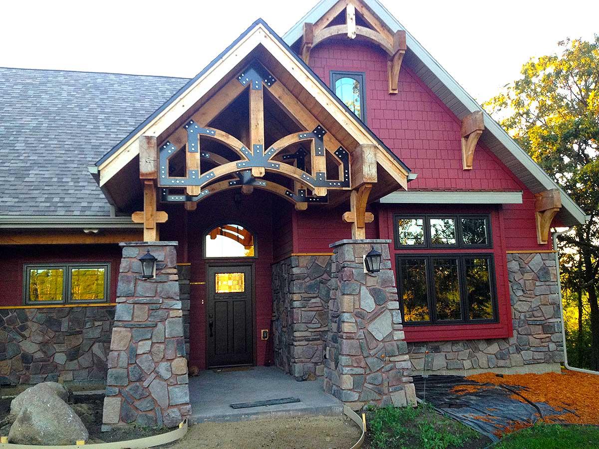 House plan exterior: Stone base, timber frame entrance, red shingle siding, and arched gable.