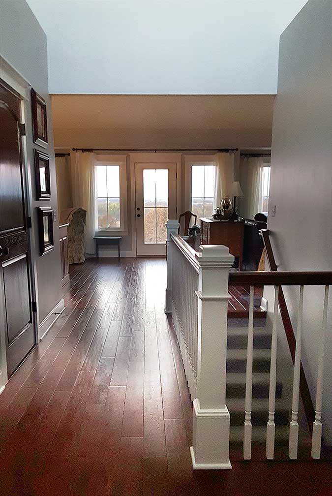 Interior view of a house hallway with hardwood floors, a staircase with white railing, and double doors leading to an exterior.