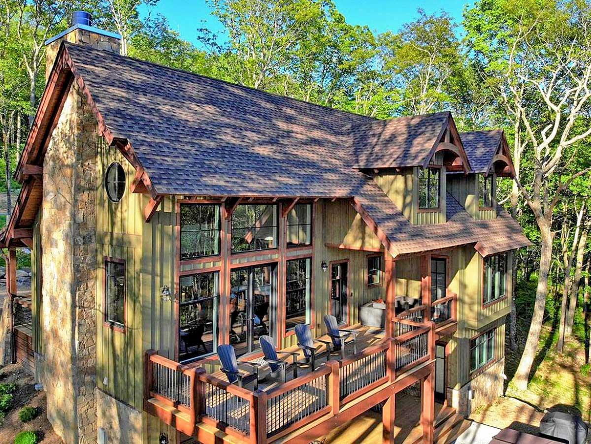 Rustic mountain house plan exterior with stone chimney, large windows, multiple gables, and expansive wrap-around deck.