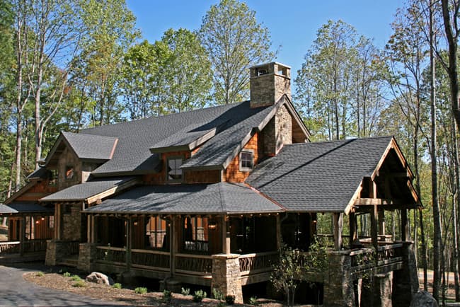 House plan exterior shows a multi-story structure with a stone chimney, gabled rooflines, dormers, and a wraparound porch with timber supports.