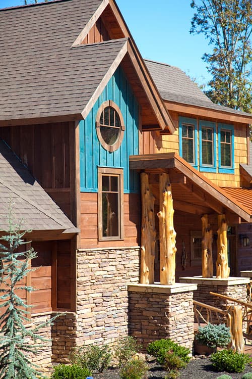 House plan exterior featuring wood siding, stone base, gables, and log posts supporting a covered entrance.