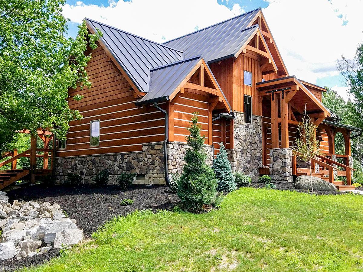 Log home exterior with stone foundation, metal roof, exposed timber framing, and covered porch with wooden railings.