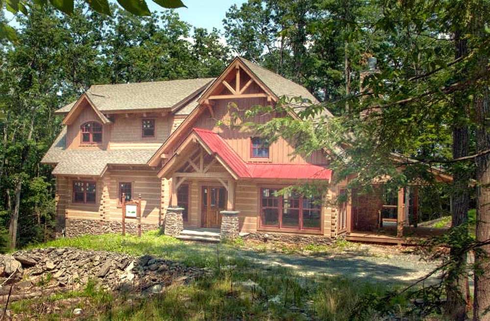 Log home exterior with a red metal roof, prominent gable with exposed timber framing, and stone-clad porch supports.