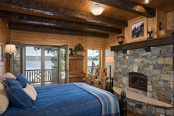 Rustic bedroom with stone fireplace, exposed wood beams, and double doors opening to a scenic view.