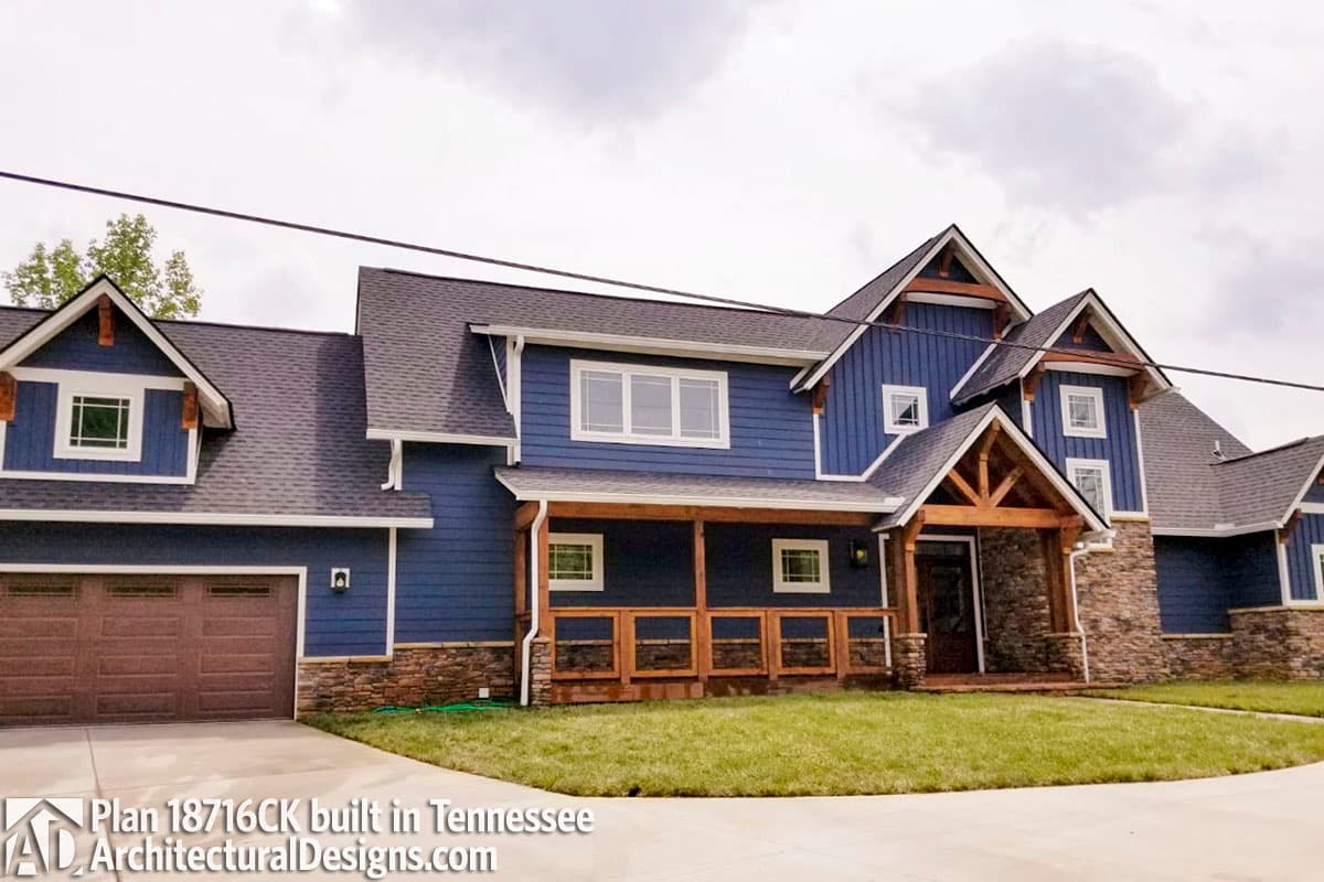 House plan exterior: two-story blue home with prominent gables, stone accents, and a covered front porch with timber supports.