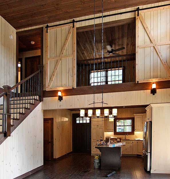 Interior view of a large room with a loft overlooking a kitchen, featuring wood paneling, barn doors, and a staircase.