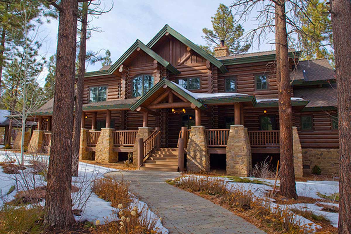 House plan exterior. Two-story log cabin with a covered porch, stone columns, and multiple gables. A walkway leads to the front door.