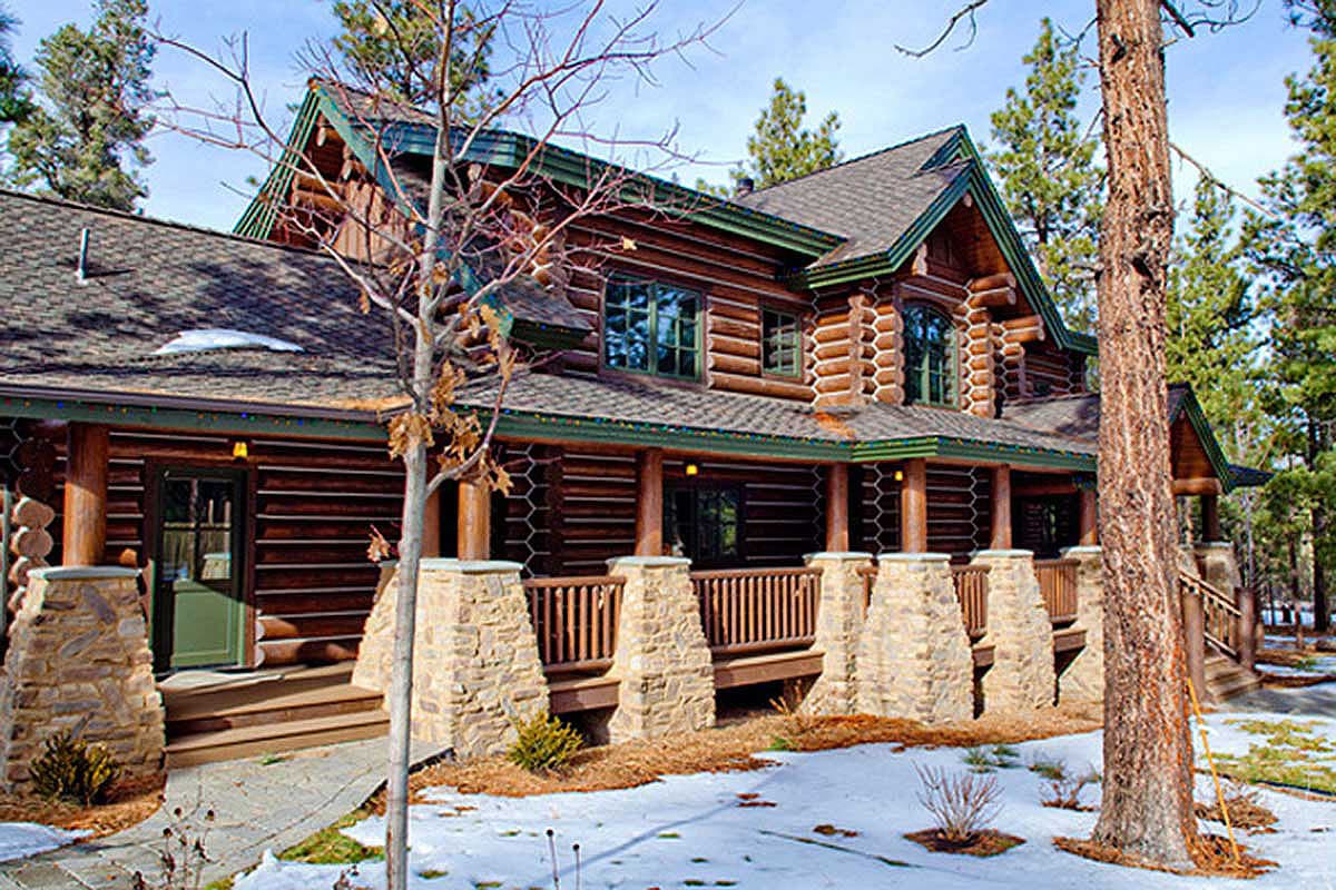 Log house exterior with a covered porch supported by stone pillars, multiple gables, and dark green trim.