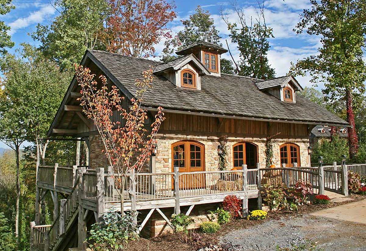 House plan exterior featuring stone and wood facade, multiple dormers, and a wraparound deck with rustic railings.