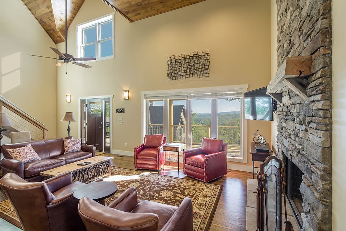 Great room with vaulted wood-beamed ceiling, stone fireplace, large windows, and leather seating.