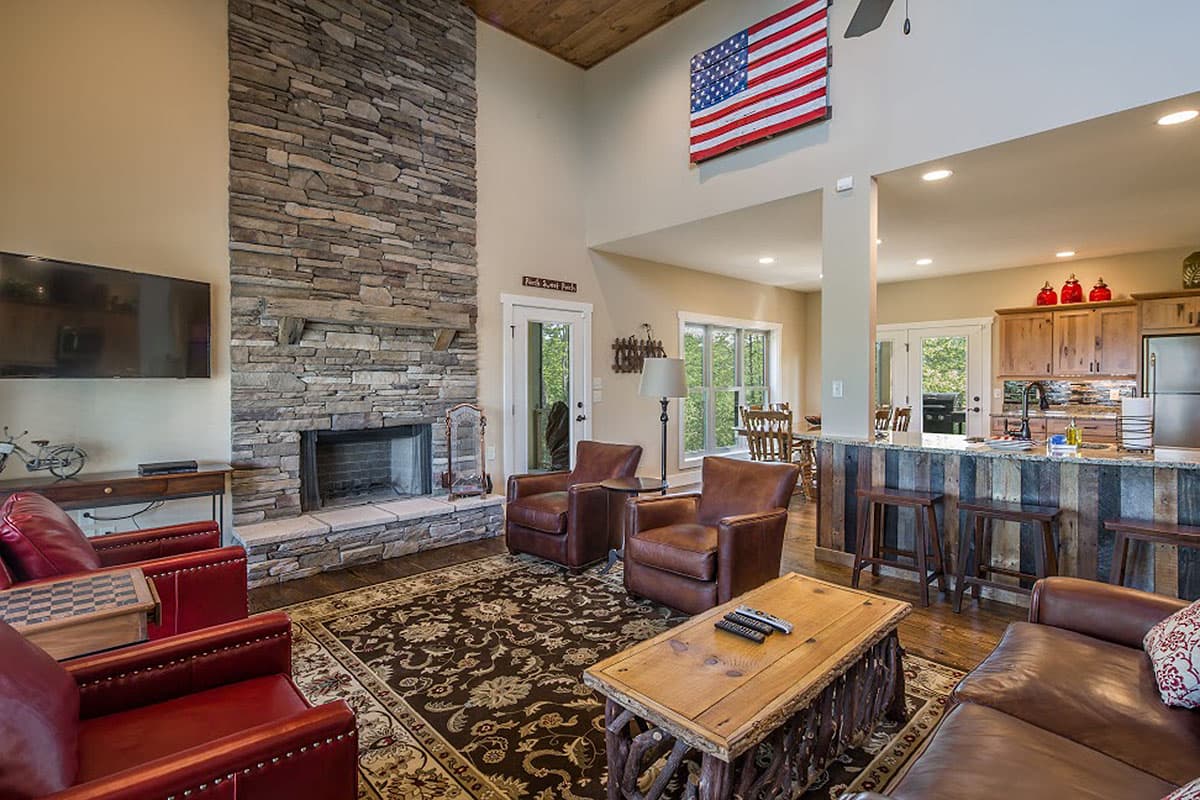 Great room with vaulted wood-beamed ceiling, stone fireplace, open kitchen with island, and dining area.