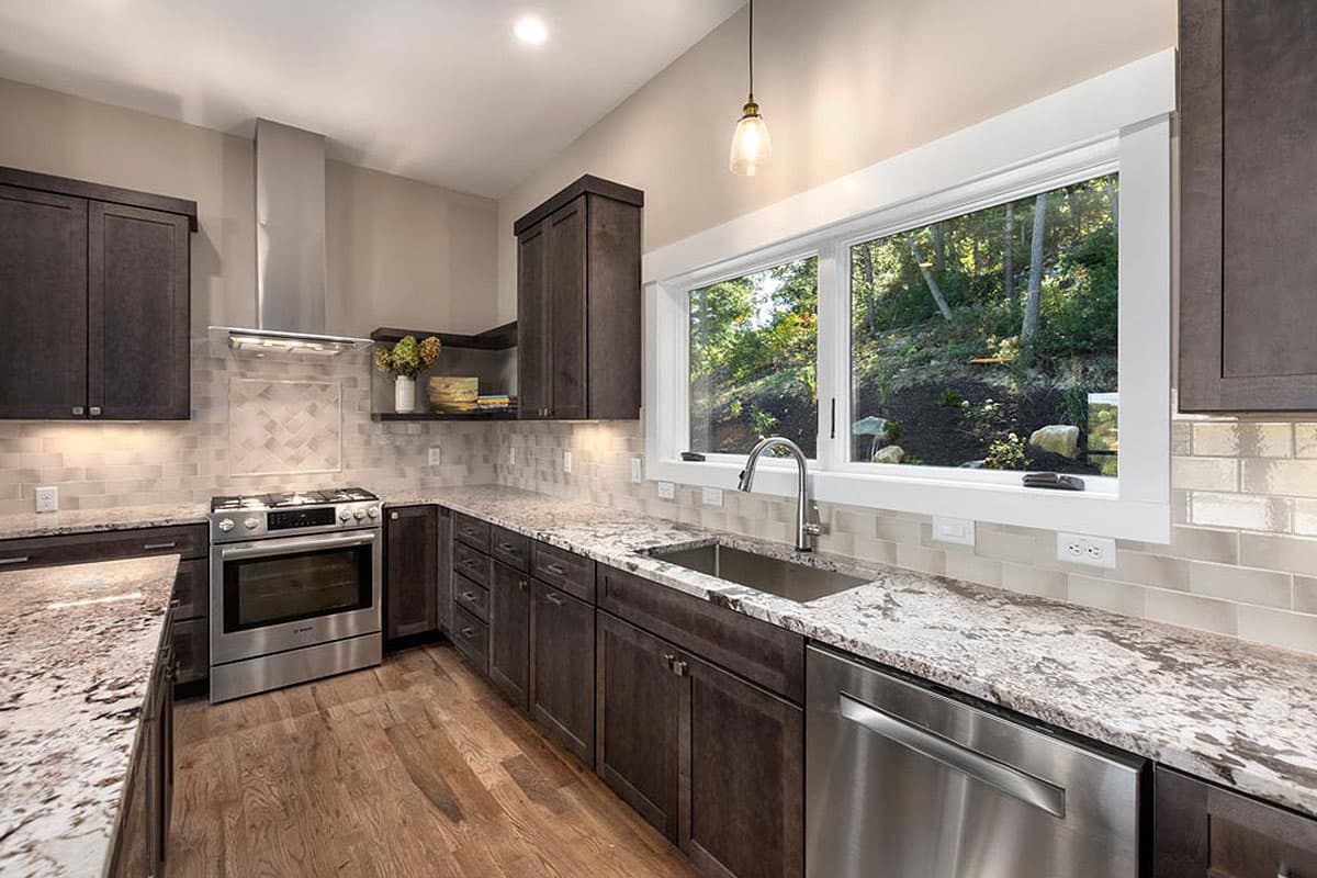 Kitchen interior with dark cabinets, granite countertops, stainless steel appliances, and large window overlooking wooded area.