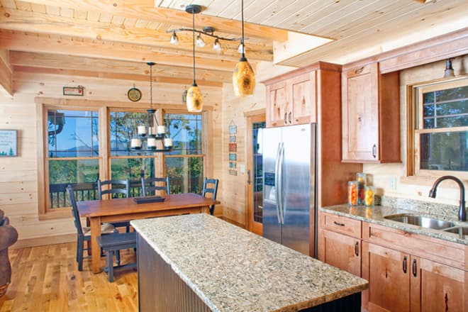 Interior view of a log cabin kitchen with a granite-topped island, stainless steel refrigerator, and dining area.