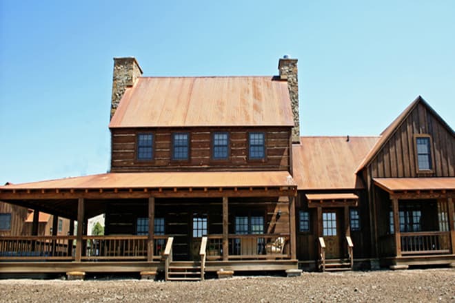 Log home exterior with a wrap-around porch, metal roof, and stone chimneys. Two stories visible.