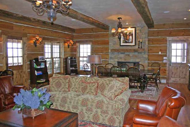 Log home living area with stone fireplace, dining table, leather armchairs, and patterned sofa. Exposed beams.