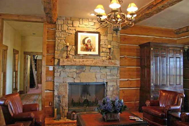Interior view of a rustic great room with a stone fireplace, log walls, exposed beams, chandelier, and wood cabinetry.
