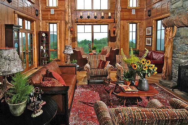 Interior view of a rustic living room with log walls, stone fireplace, leather sofa, patterned armchairs, and large windows with mountain views.