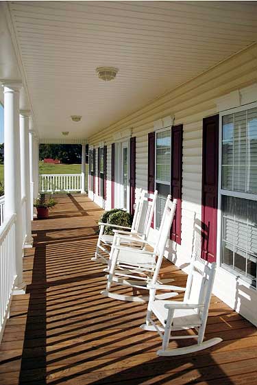 House plan exterior with a covered porch featuring white columns, white railings, wood decking, and multiple rocking chairs.