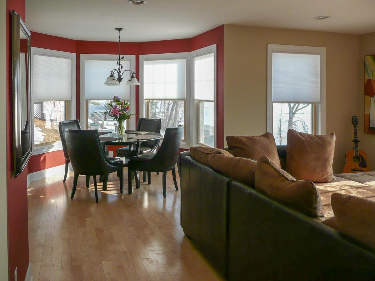 Interior view of a dining area with a round table, four chairs, and bay windows, adjacent to a living area with a sectional sofa and guitar.
