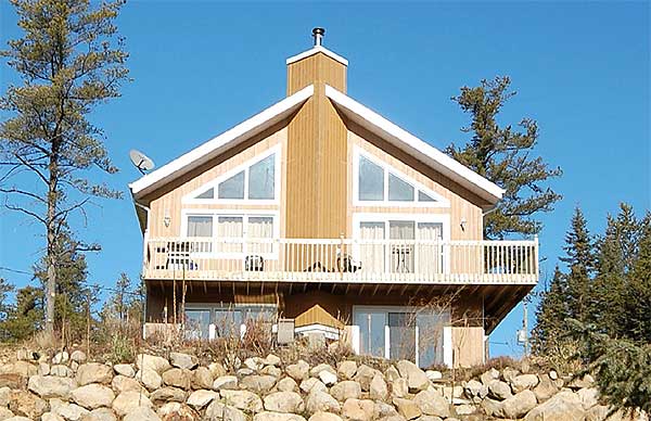 A-frame cabin with large triangular windows, a balcony, and a stone retaining wall in front. Trees surround the house, and a blue sky is visible.