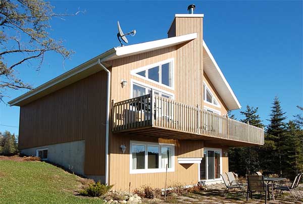 A-frame house with a balcony, wooden siding, and large windows. The house has a satellite dish on the roof and is surrounded by green lawn and trees.
