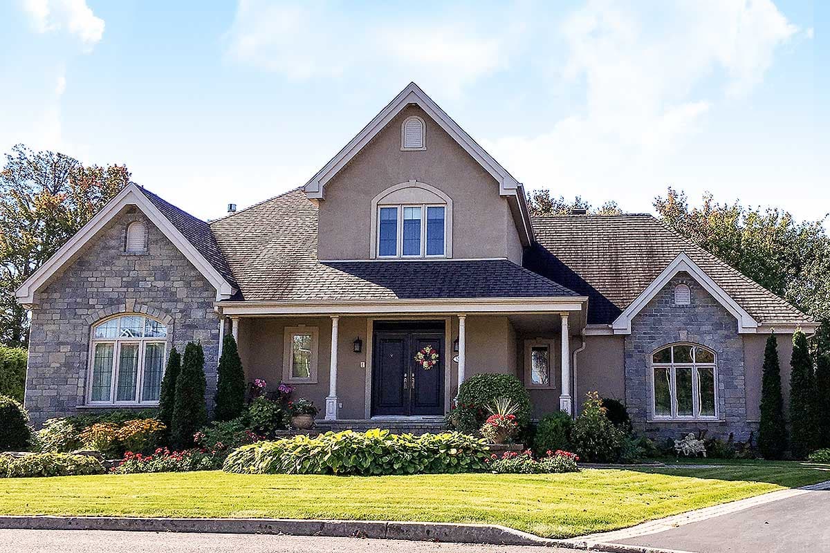 House plan exterior. Two-story, stucco and stone facade. Features include a front porch with columns, multiple gables, and a dark shingle roof.