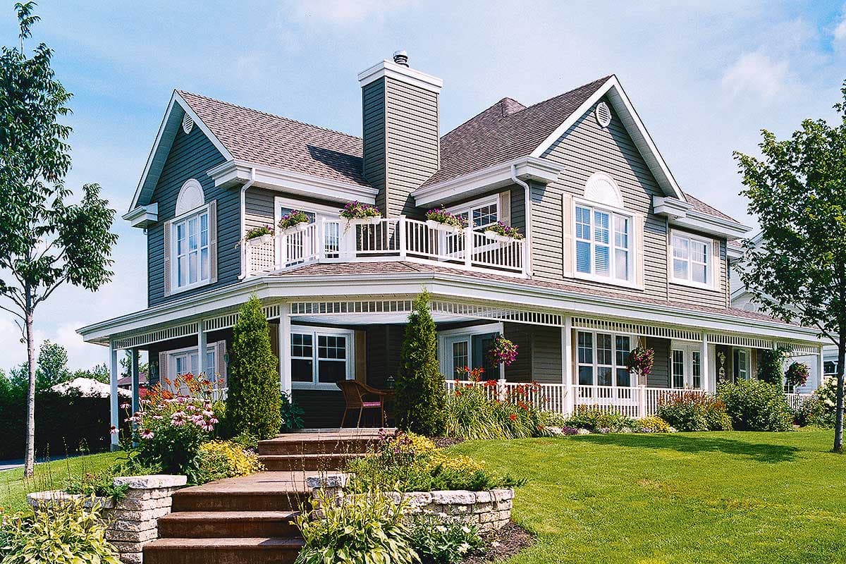 House plan exterior: Two-story home with wraparound porch, second-story balcony, gables, dormers, and prominent chimney.