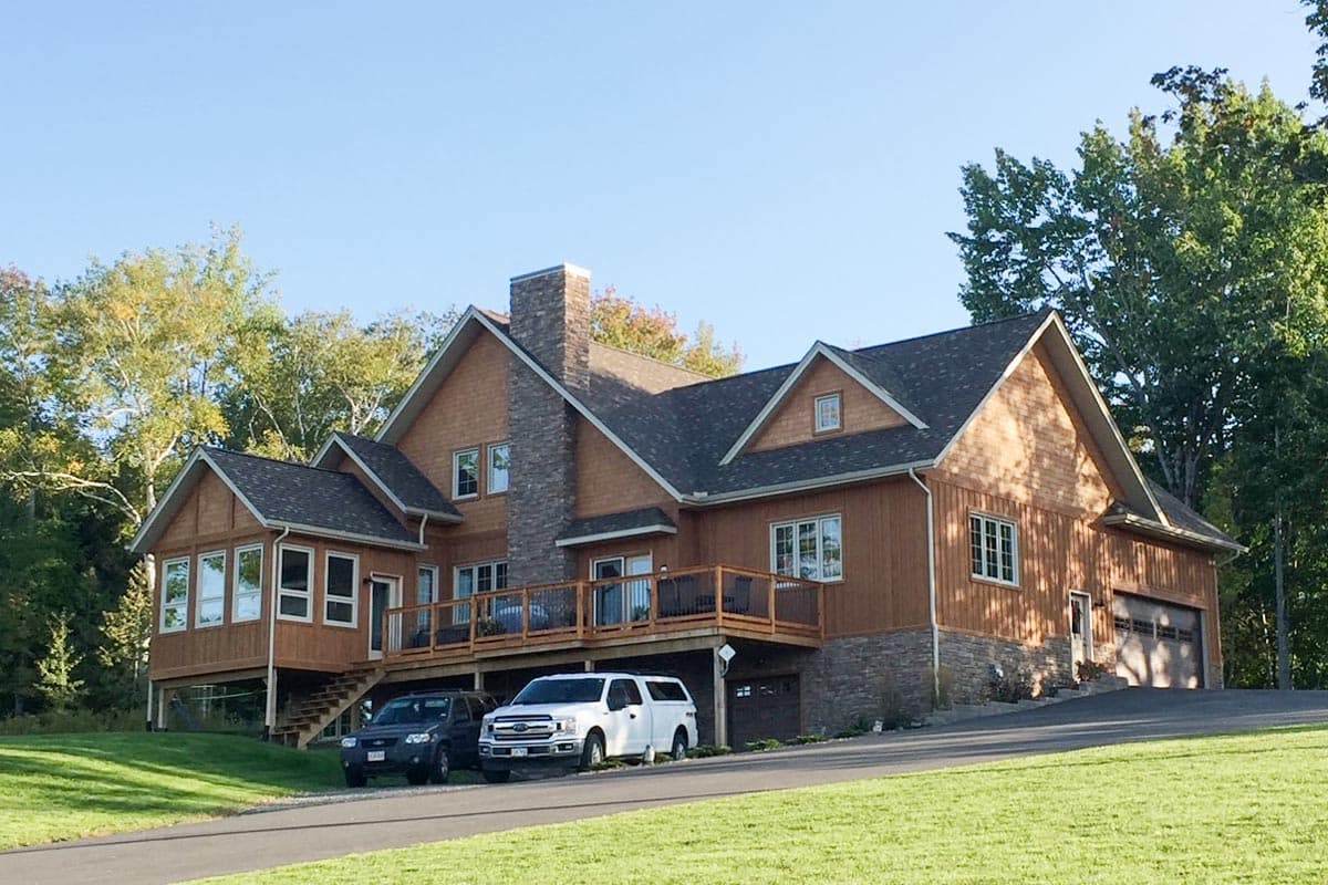 House plan exterior with wood siding, stone foundation, large deck, and attached garage. Features multiple gables and a prominent stone chimney.