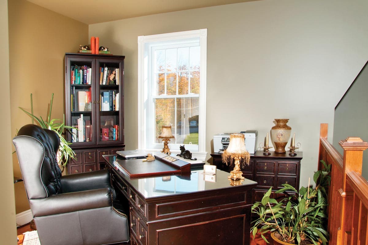 Home office interior with a desk, tufted leather chair, bookcase, printer, and a window overlooking autumn trees.