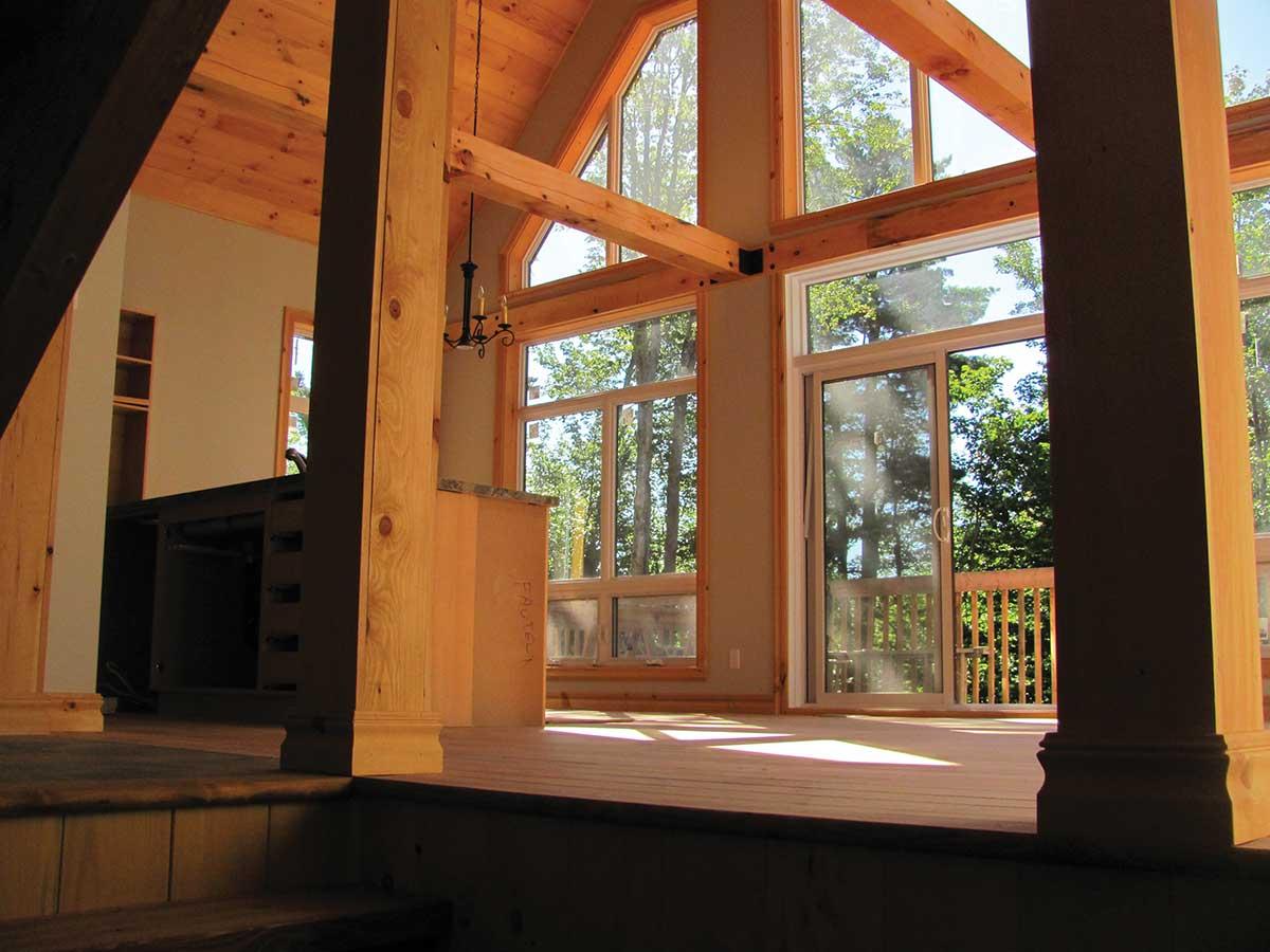 Interior view of a timber-framed house with vaulted ceilings, exposed beams, and large windows looking out to trees.