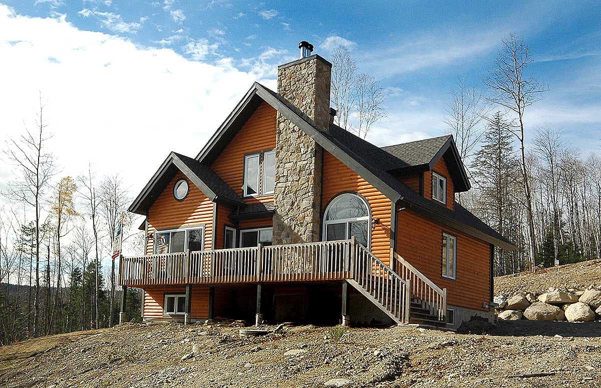House plan exterior: Log home with stone chimney, wraparound deck, arched window, and multiple gables.