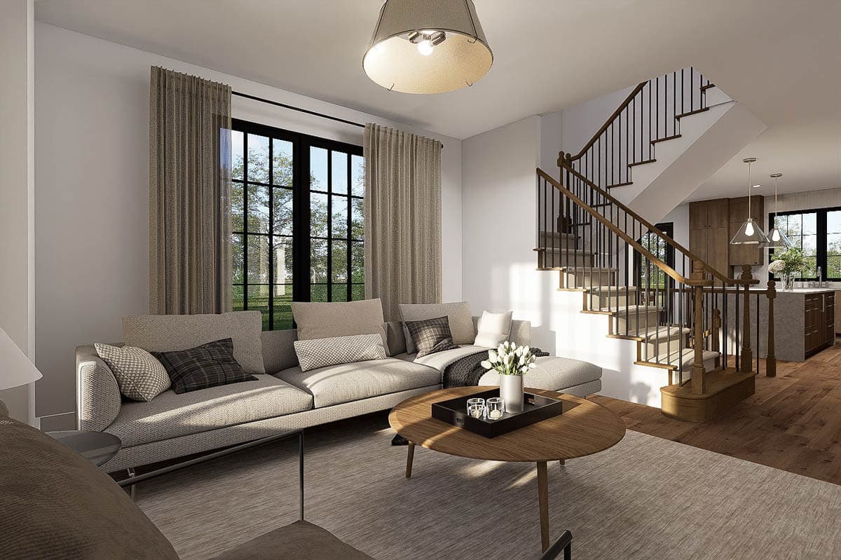 A modern living room with a neutral color scheme. A large sectional sofa faces a round coffee table. A staircase and kitchen are visible.