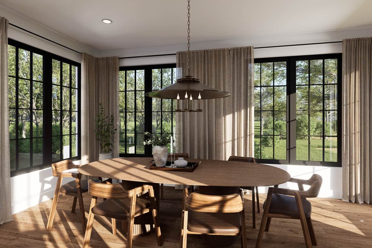 A sunlit dining room features a large oval wooden table with six chairs. A unique chandelier hangs above the table, and large windows offer a view.