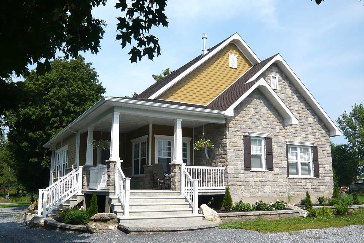 House plan exterior. Two-story with stone facade, covered porch, and gabled roof. Features include white columns, dark shutters, and dormer windows.
