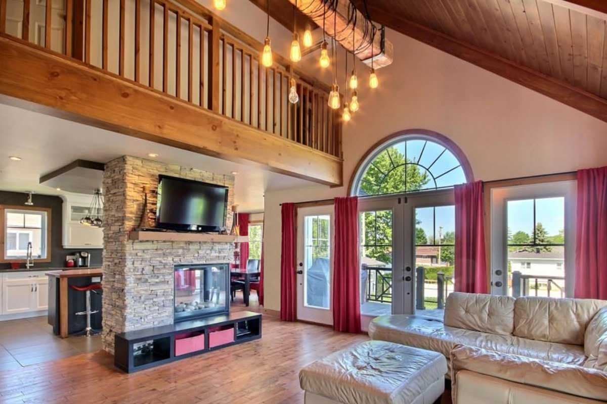 Living room interior with stone fireplace, open loft, vaulted ceiling, and large arched window.