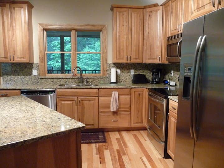 Kitchen with natural wood cabinets, granite countertops, stainless steel appliances, and a double-hung window overlooking trees.