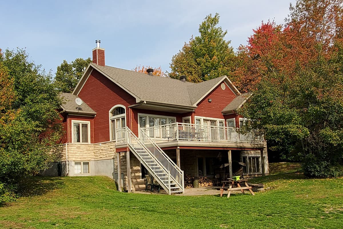 House plan exterior view of a two-story red house with stone foundation, large deck with stairs, and gabled roof.