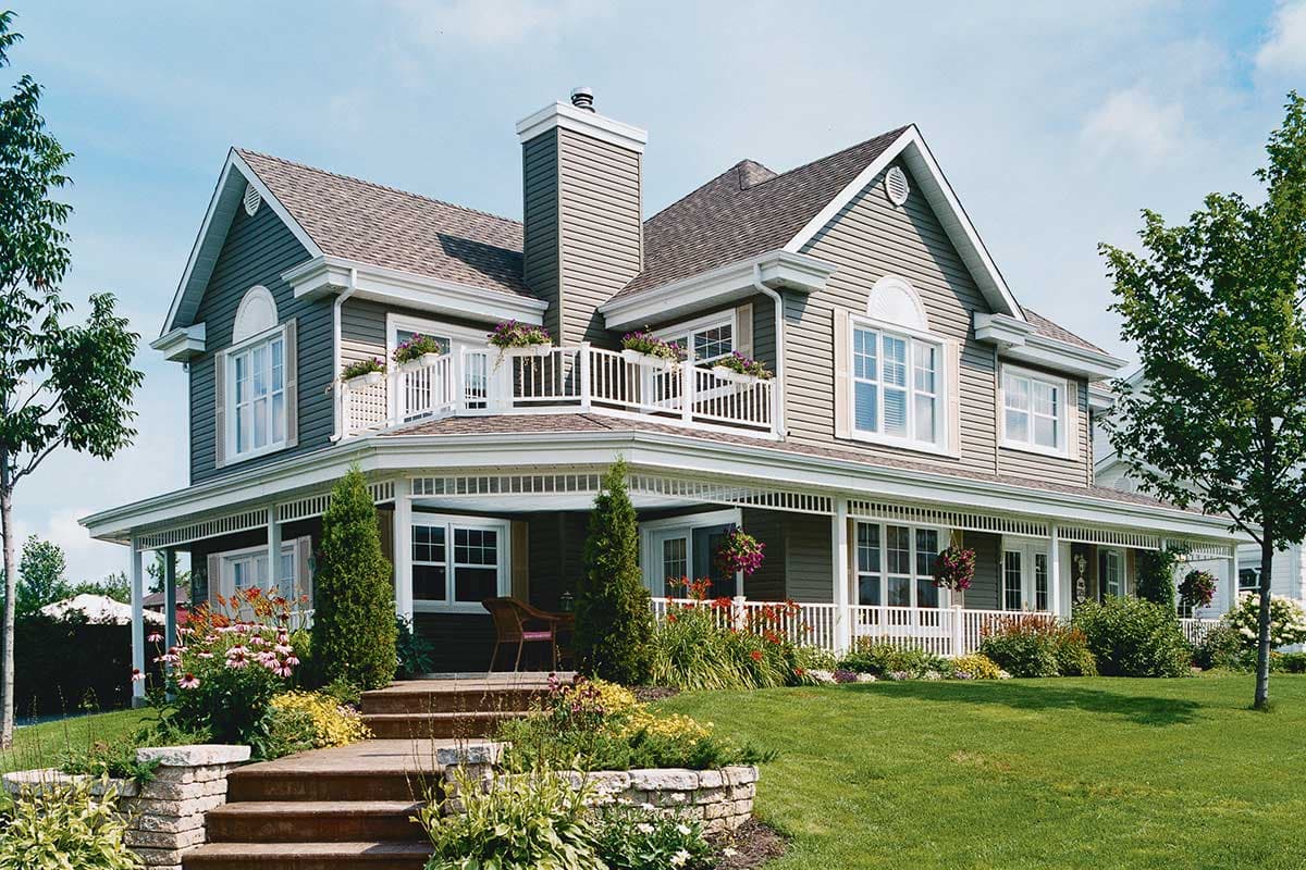 House plan exterior: two-story home with wraparound porch, second-story balcony, dormers, and prominent chimney.