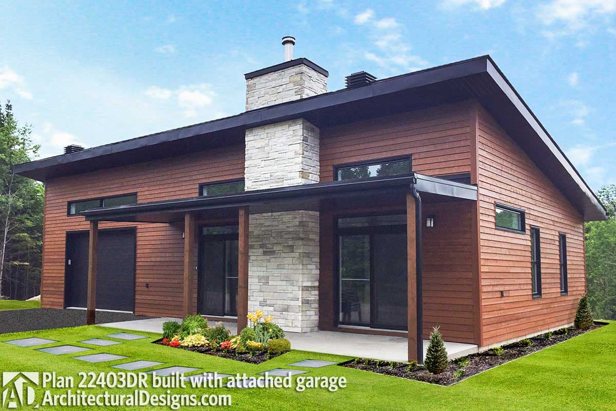Modern, wood-paneled home with a stone chimney, attached garage, and a covered porch. Lush green lawn with stepping stones and flower beds.