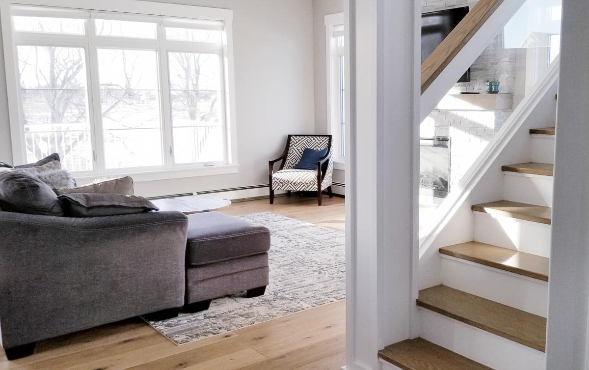 Interior view of a living room with large windows, sectional sofa, accent chair, and a staircase with glass railing.