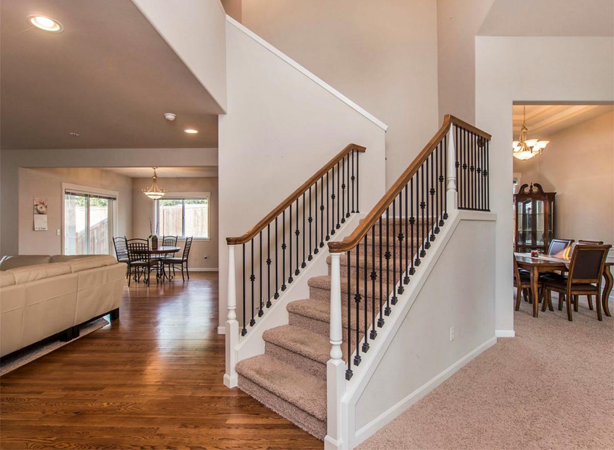 Interior view of a house with a carpeted staircase featuring dark spindles and wooden handrails. Adjacent dining and living areas visible.