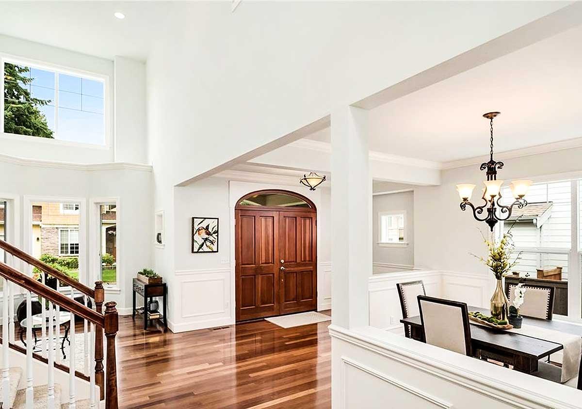 Interior view of a two-story foyer with double front doors, a grand staircase, large windows, and a dining area.