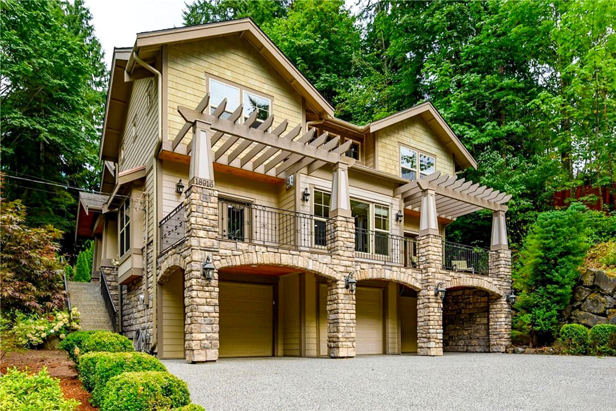 House plan exterior with two stories. Features include a stone facade, pergola-covered balcony, and arched garage entrances. Shingle roof.